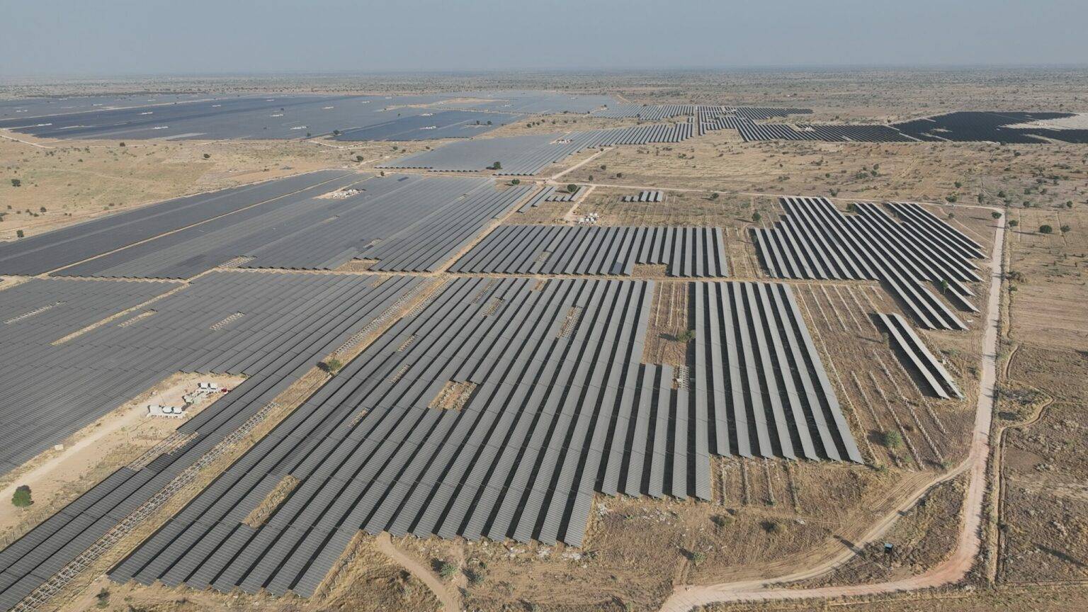 Aerial view of large solar panel arrays arranged in geometric rows across an arid landscape, with a highway running alongside and water retention ponds visible in the distance.