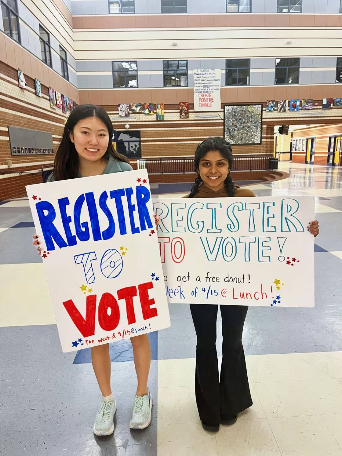 Two high school students hold signs that say "Register to Vote" in a large room in their school