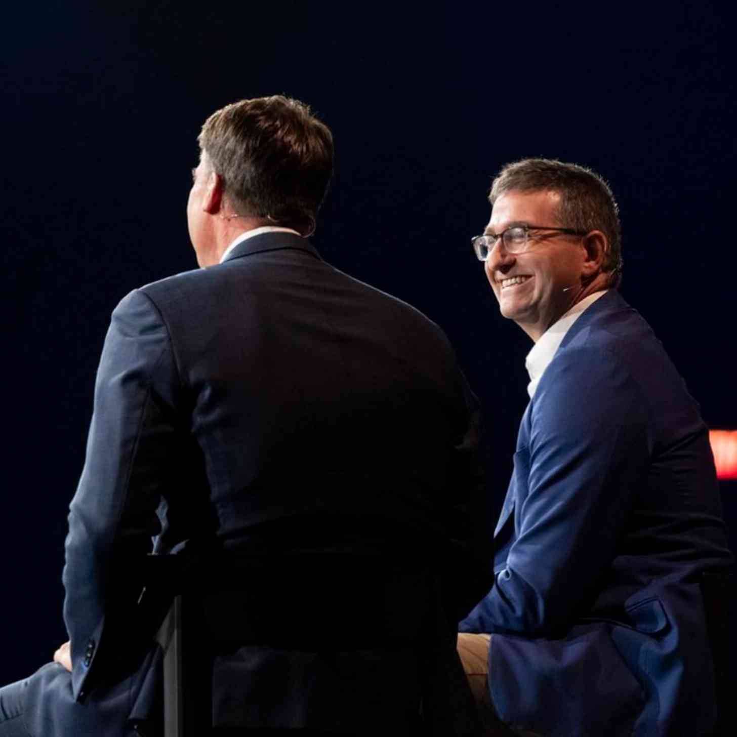 Two lawmakers seen from behind wearing microphones on chairs on a stage