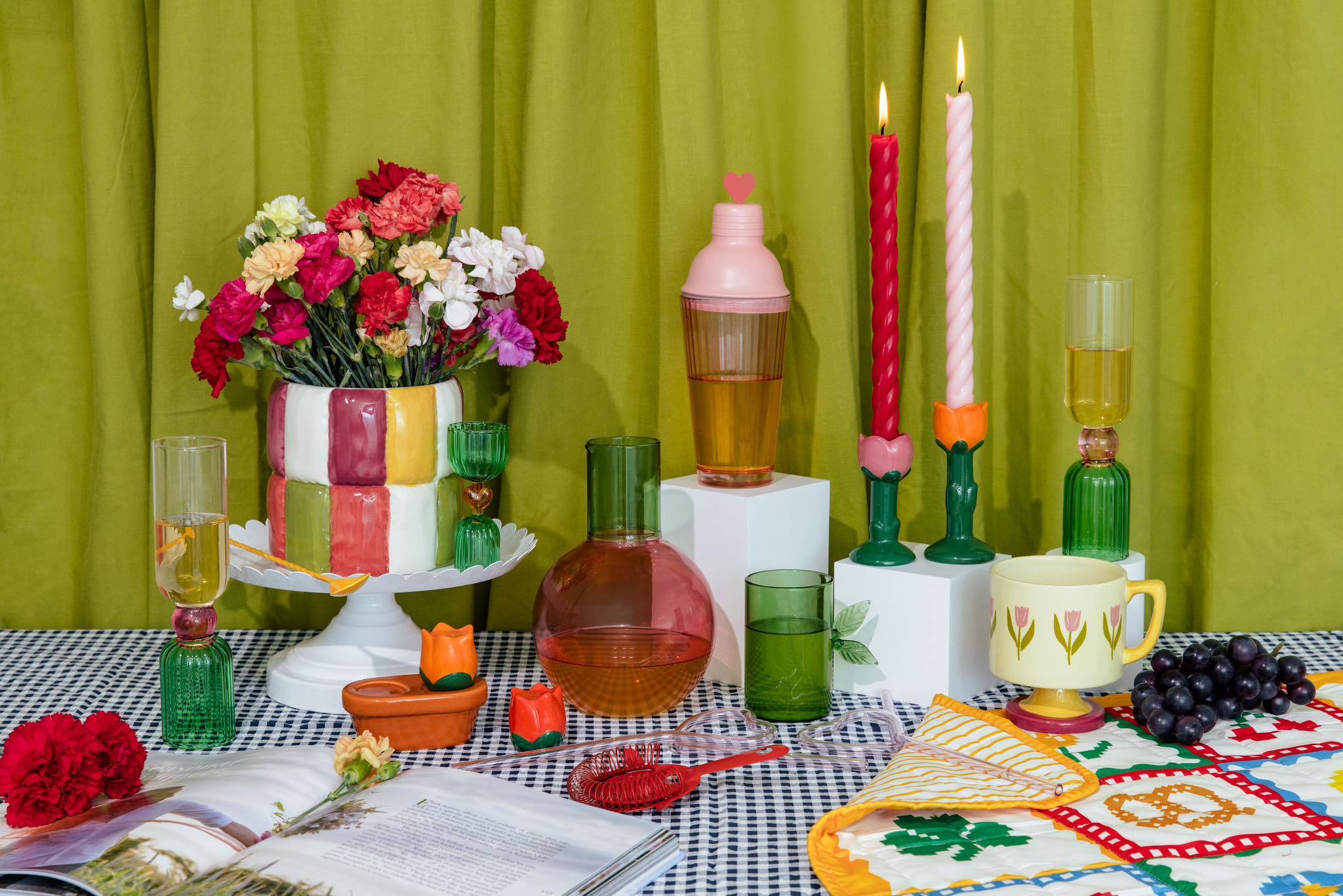 Green, red, and orange-colored champagn glasses, vases, decanters, glasses, and more on a plaid tablecloth against a green curtain background