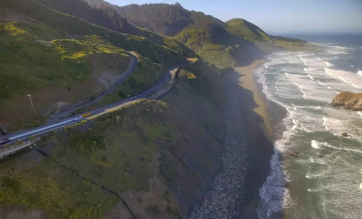 Aerial photo of a two-lane highway in the side of a cliffside next to waves on a beach