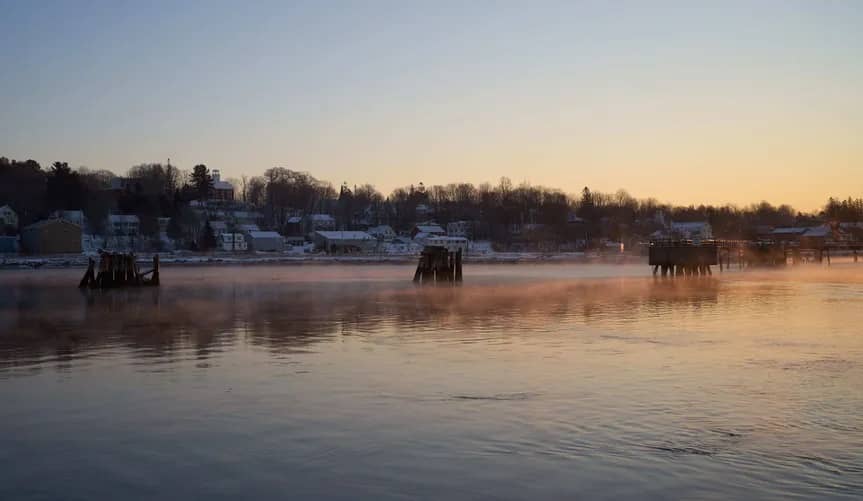 Taken from over a body of water looking back at houses along the shoreline, mist rises from the top of the water at sunrise