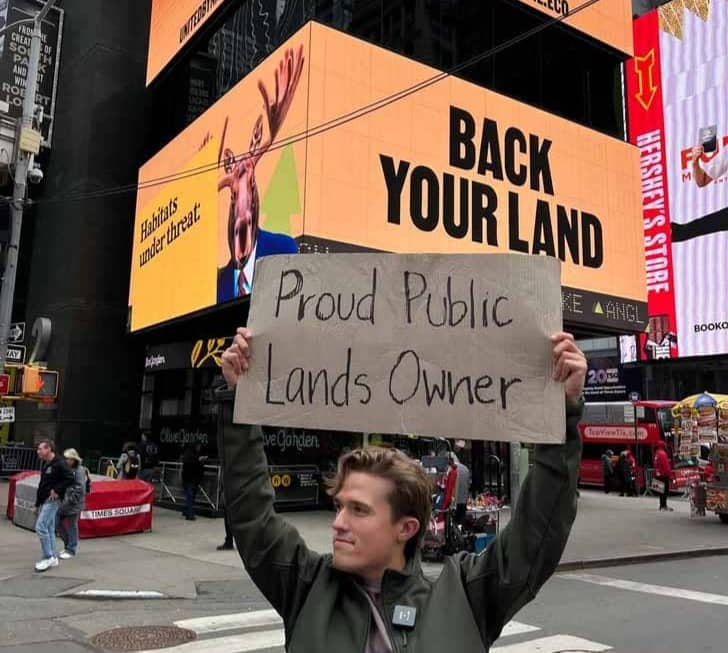 Person holding a cardboard sign that says "Proud Public Lands Owner" in front of a Times Square billboard that says "Back Your Land"