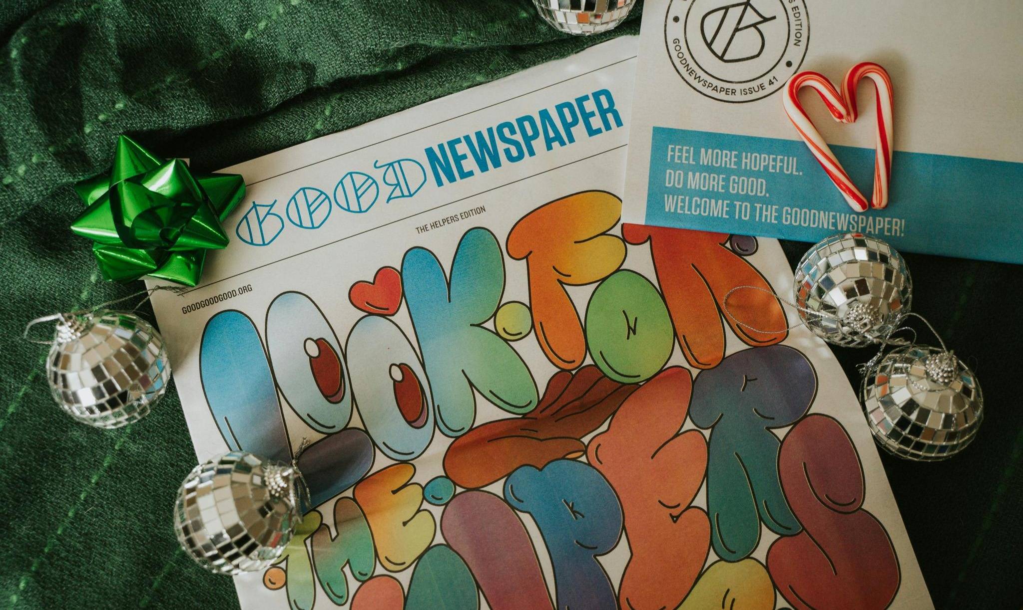 A Goodnewspaper laying on green fabric, it has a green bow on it and is surrounded by mirrorball ornaments