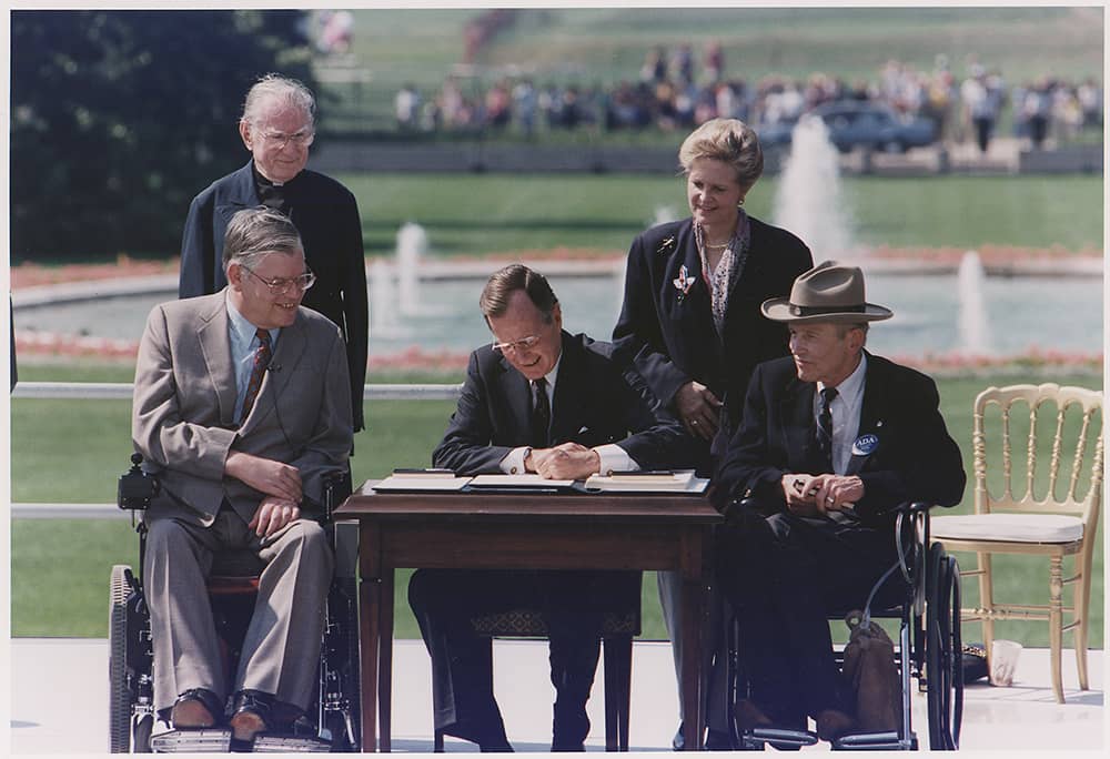 A person sitting at a small wooden desk, signing a paper, two people in wheelchairs are on either side of hi, and two people are standing in behind the three of them.