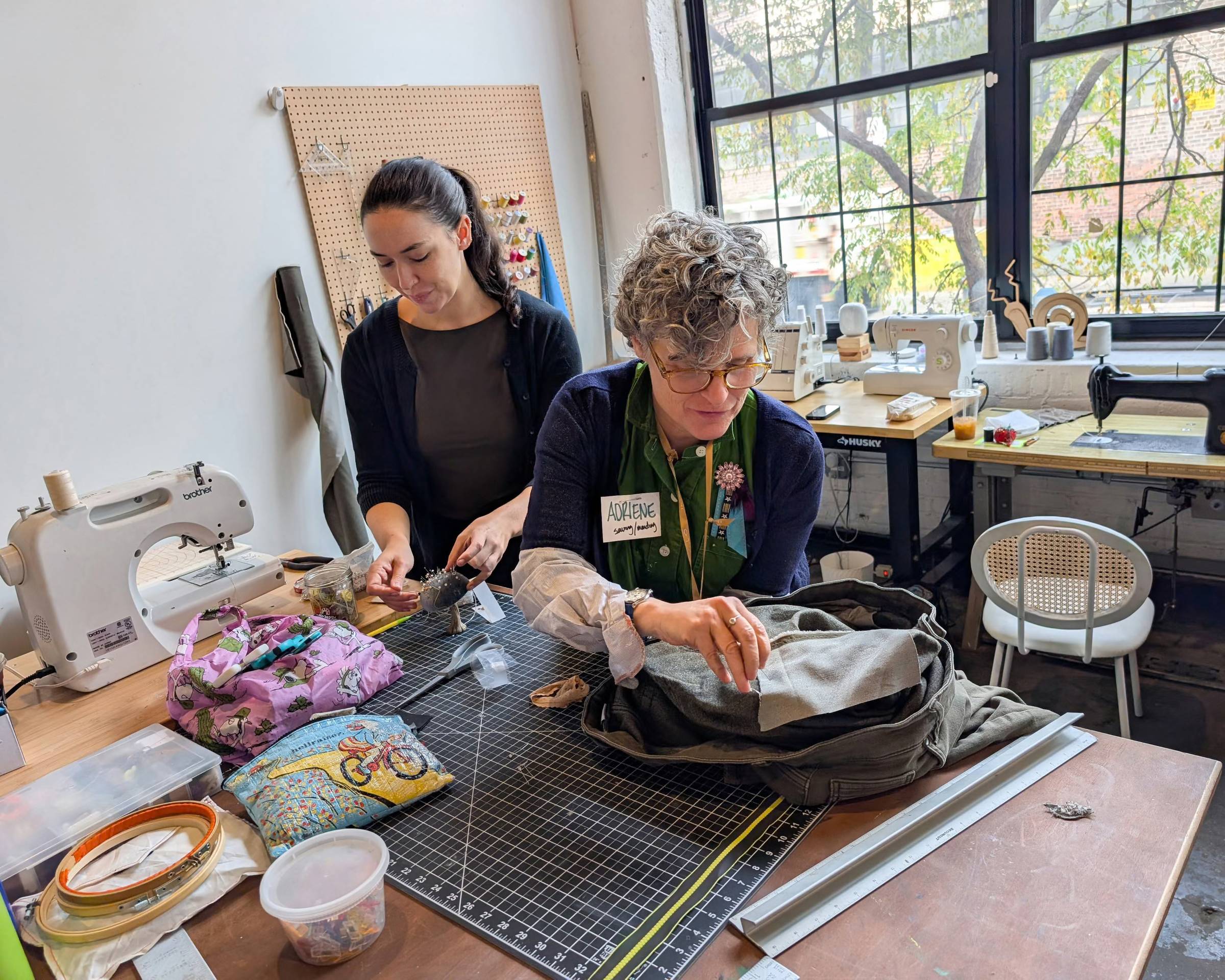 Two women work at a crafting table in a bright studio space with large windows. The older woman wearing glasses and a green apron examines fabric from an open bag, while the younger woman stands beside her. A sewing machine, embroidery hoops, colorful fabric, and crafting supplies are visible on the work surface.
