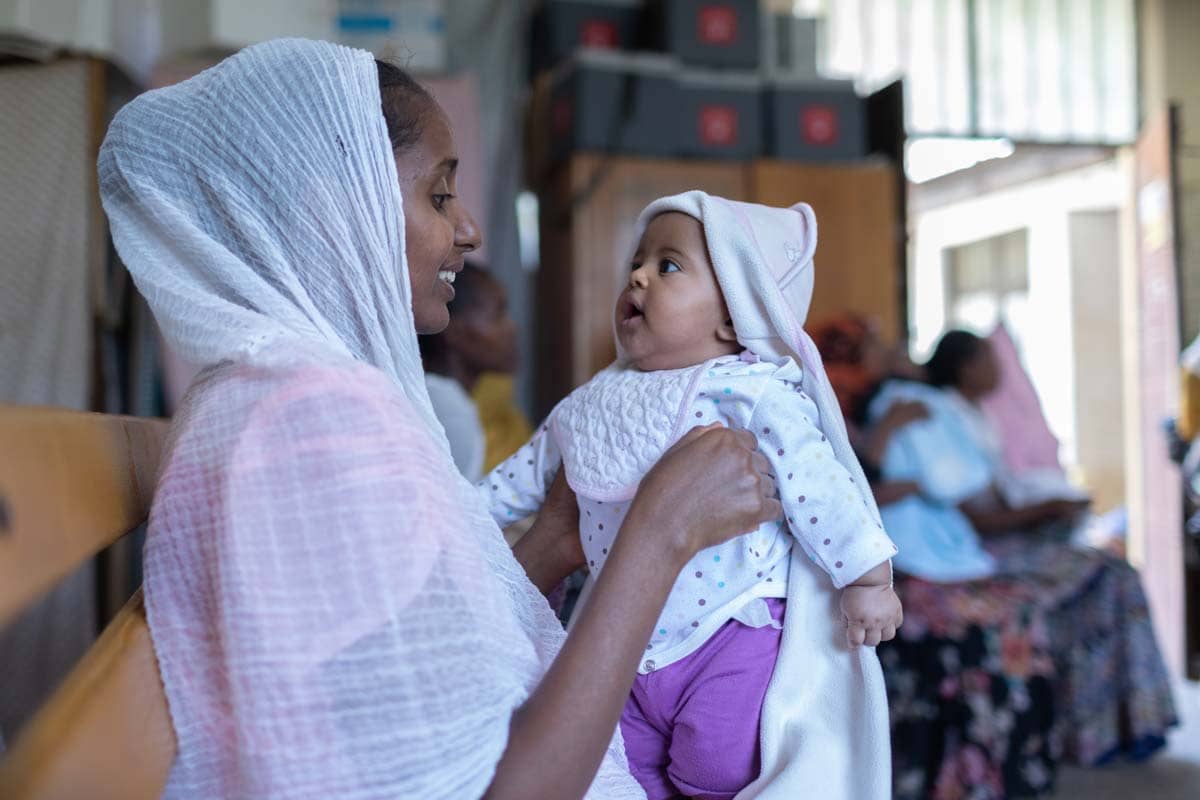 A mother in a white head scarf smiling and holding a baby wearing pants, a shirt, a bib, and blanket