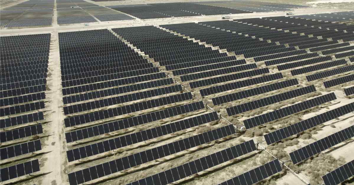 Aerial view of a large solar panel farm with rows of dark photovoltaic panels arranged in angled lines across flat terrain, separated by light-colored access roads.