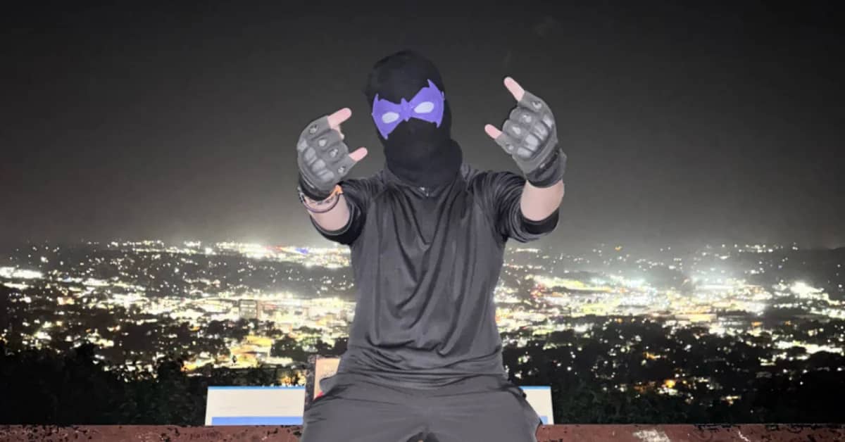 Person sitting on a wall with a lit-up city behind them, they're wearing all back and a ski mask with purple mask over the eyes