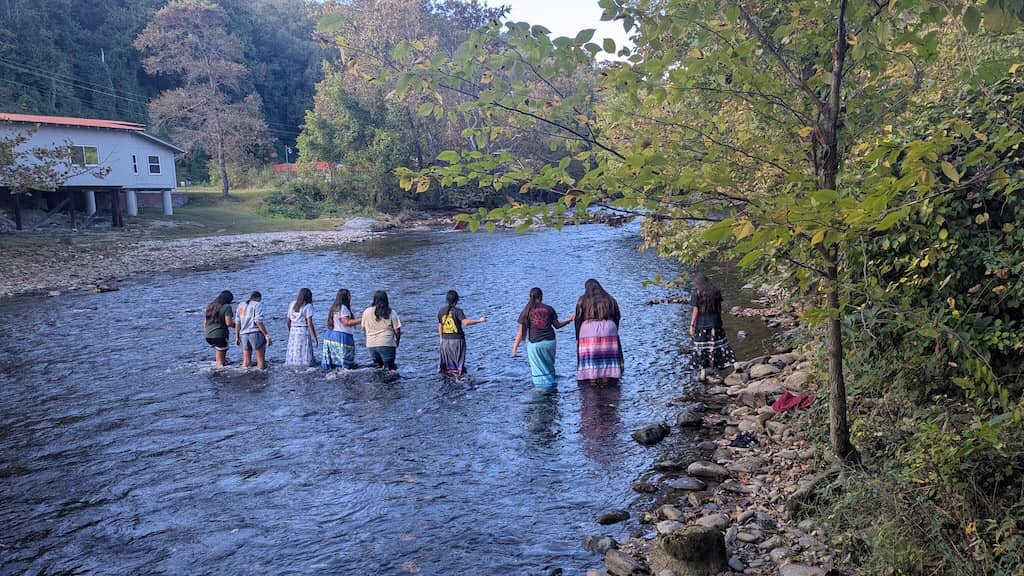 A group of about ten people stand in shallow water in a river, wearing casual clothing. Trees with green and autumn-colored foliage line both banks, and a small white house with a red roof sits on the left bank.