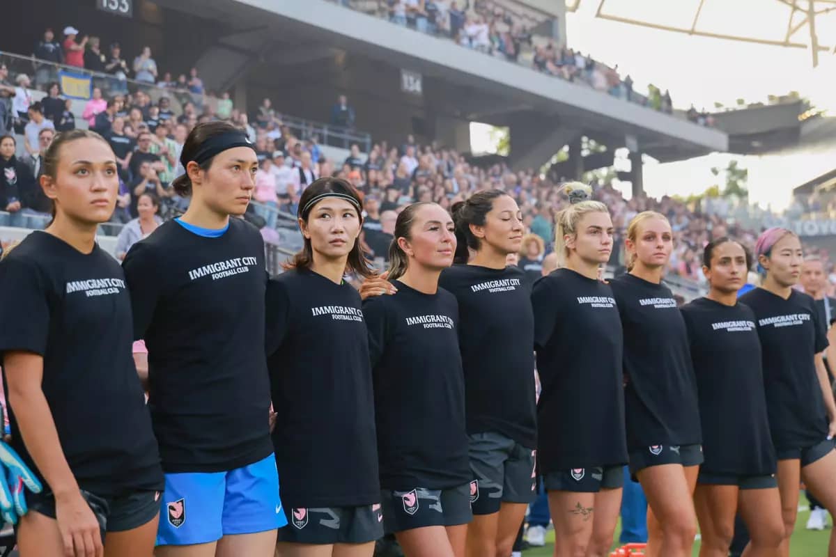 Players lined up with black shirts that say "immigrant city football club"