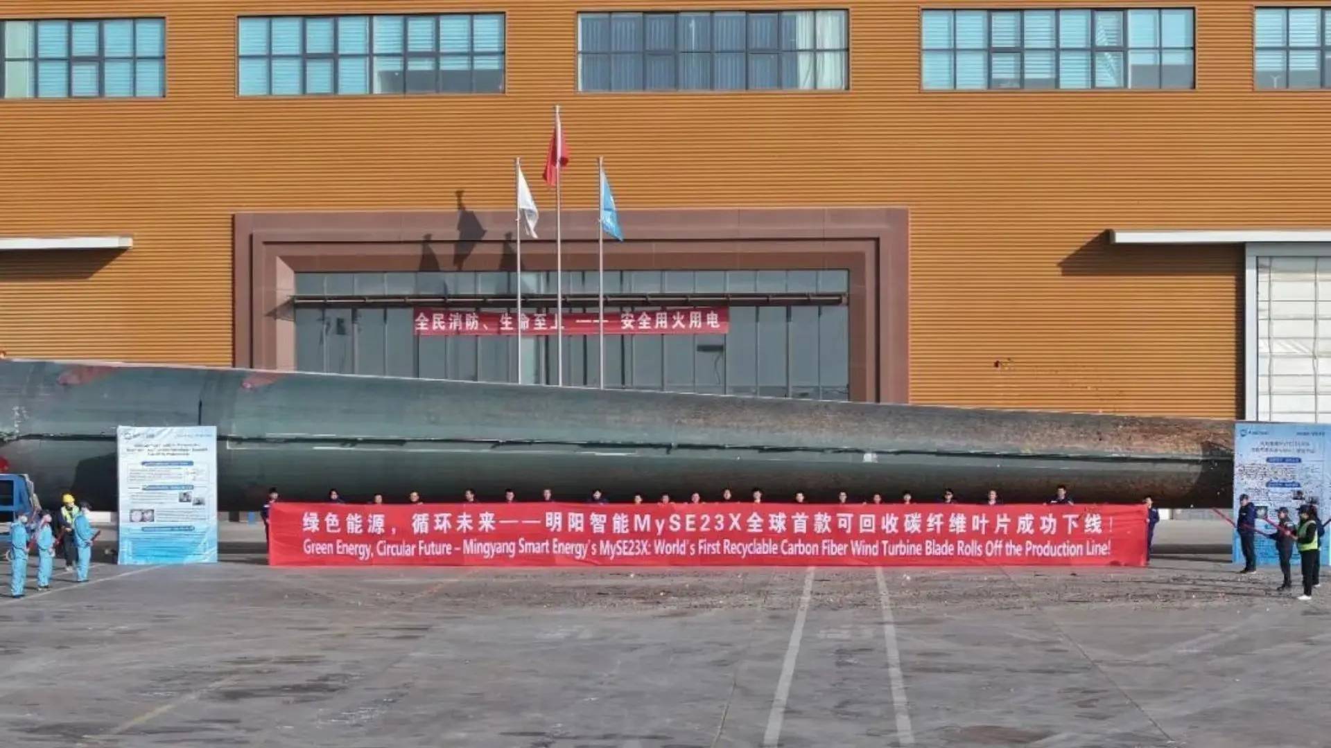 A large wind turbine blade with a red banner that reads "Green Energy, Circular Future - Mingyang Smart Energy's MySE23X: World's First Recyclable Carbon Fiber Wind Turbine Blade Rolls Off the Production Line!"