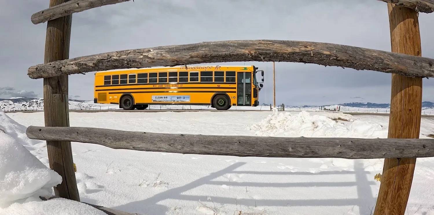 An electric yellow school bus on a road surrounded by snow with fence posts in the foreground and small mountains in the background