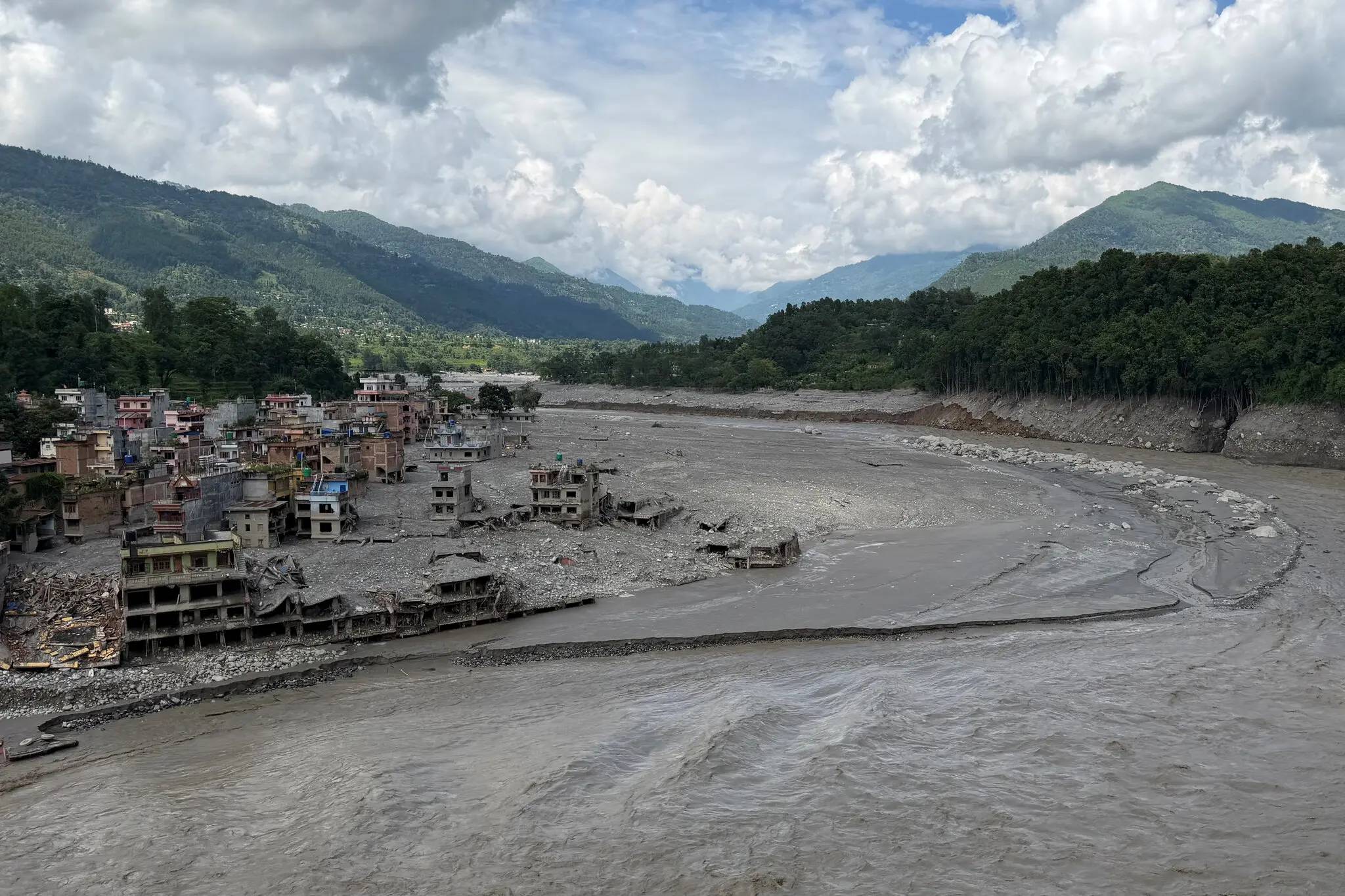 A river with murky brown water flows past a settlement of multi-story buildings on the left bank, with ruined structures visible in the riverbed. Mountains covered in green forest rise in the background under a cloudy sky.