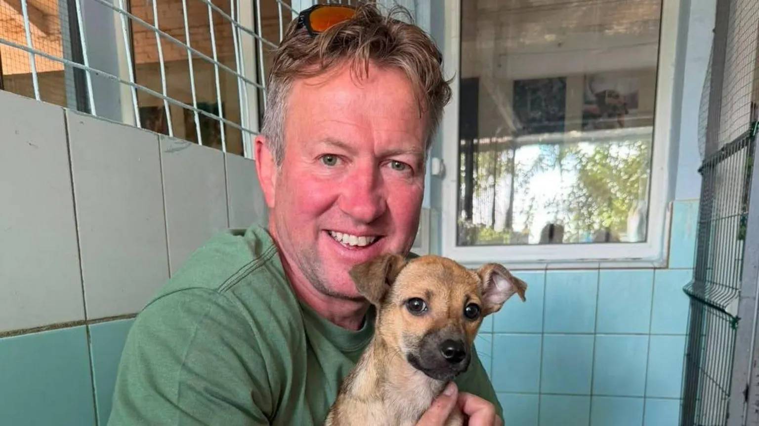 A man in a green shirt holding a small dog, they are both looking at the camera