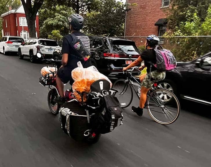 Two bikers with bulk bags of food on the back of one bicycle