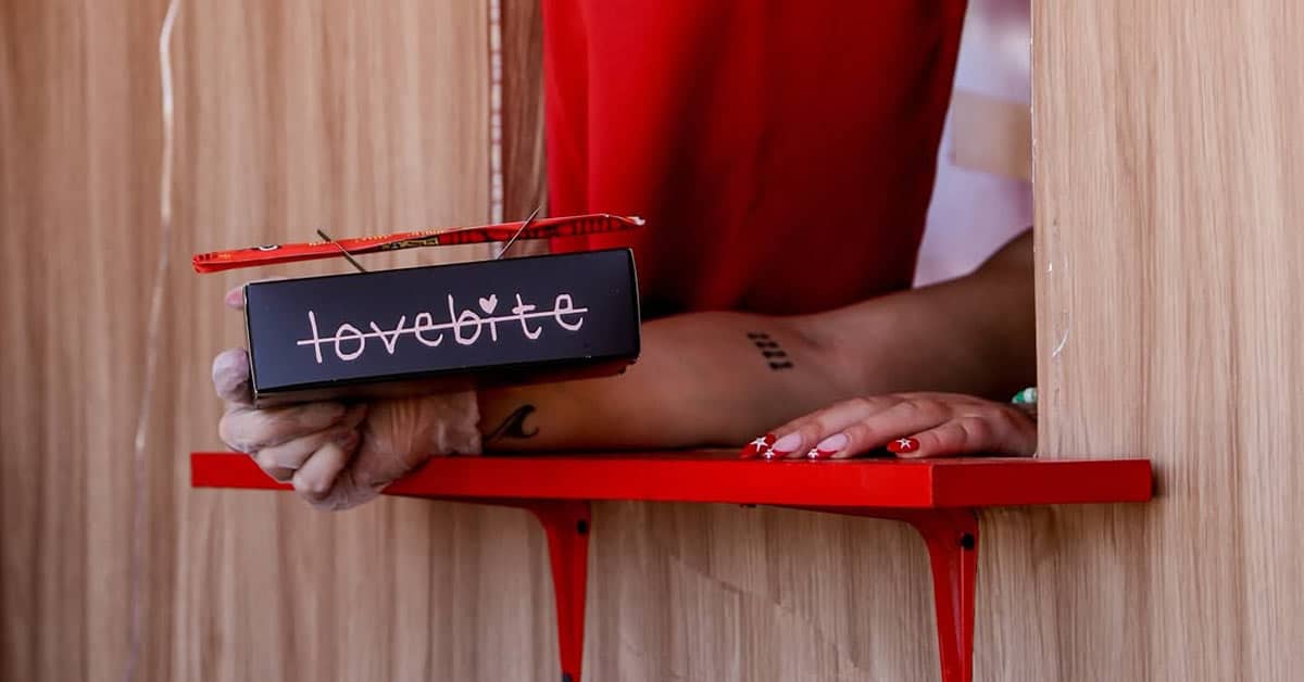 A hand holding a "lovebite" to-go box out of a delivery window with a red tray on the outside