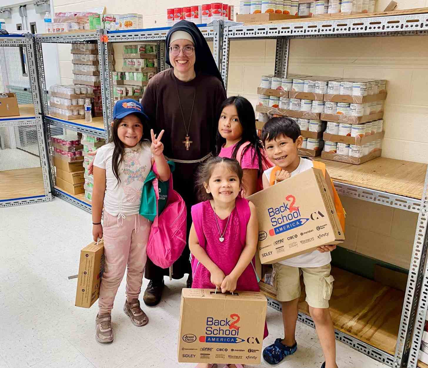 A nun standing in a food pantry with four children around her holding "back 2 school America" boxes