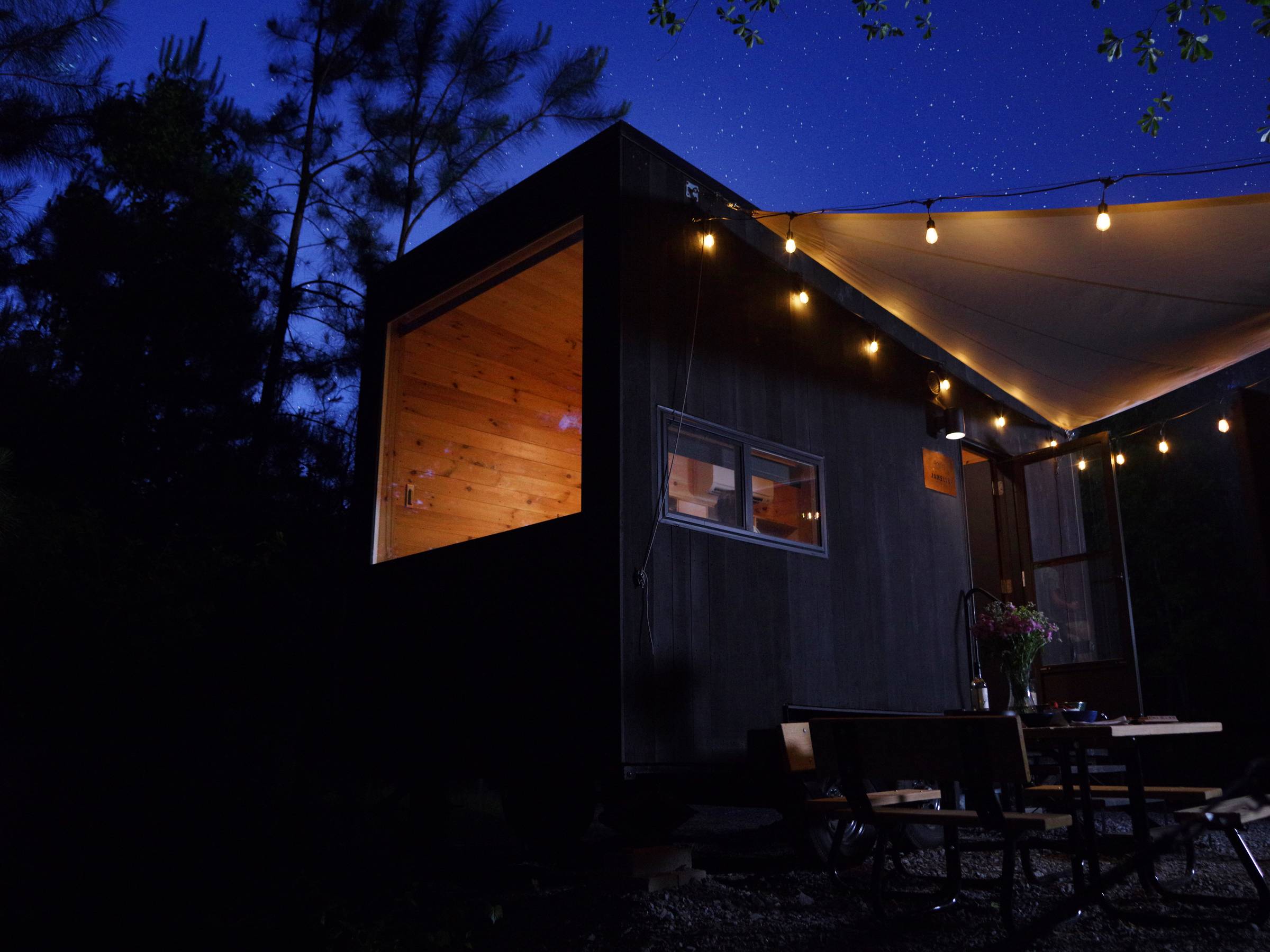 A tiny camper with a black exterior, large window on the end, canopy and string lights in the middle of a dark forest with a starry sky overhead
