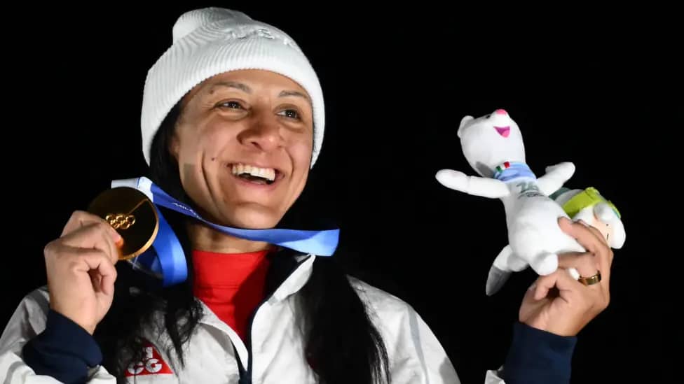 Elana Meyers Taylor in a white beanie holding up her gold medal and smiling