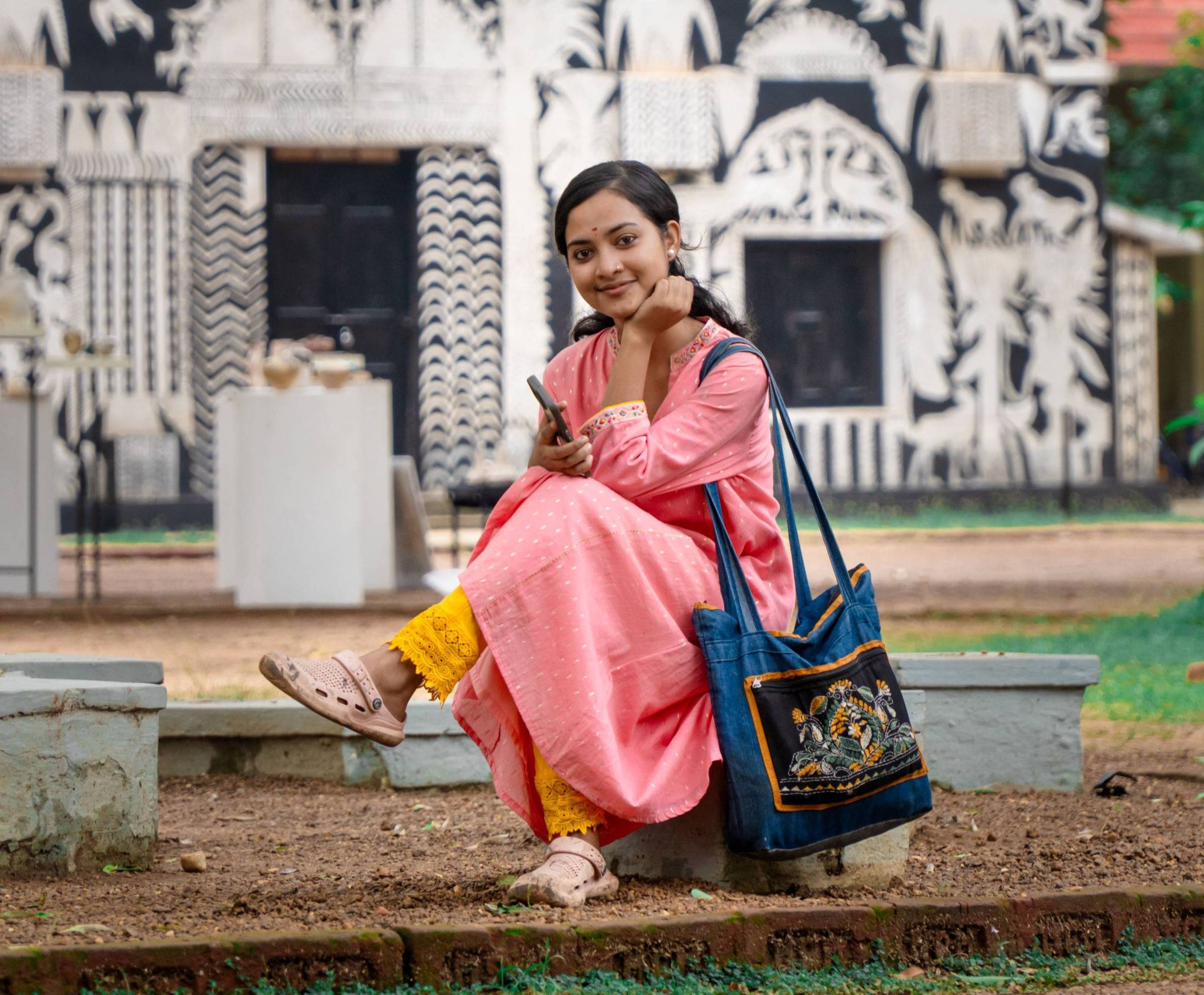 A young woman in a pink dress and blue bag sits on a concrete bench in front of an ornate white and black building with decorative architectural details and dark windows.