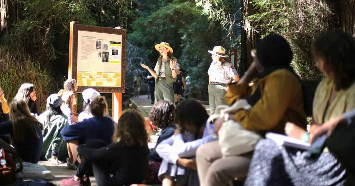 Two park rangers in uniform and hats give a presentation to a seated audience beneath tall trees. An informational sign is visible on the left side of the gathered group.