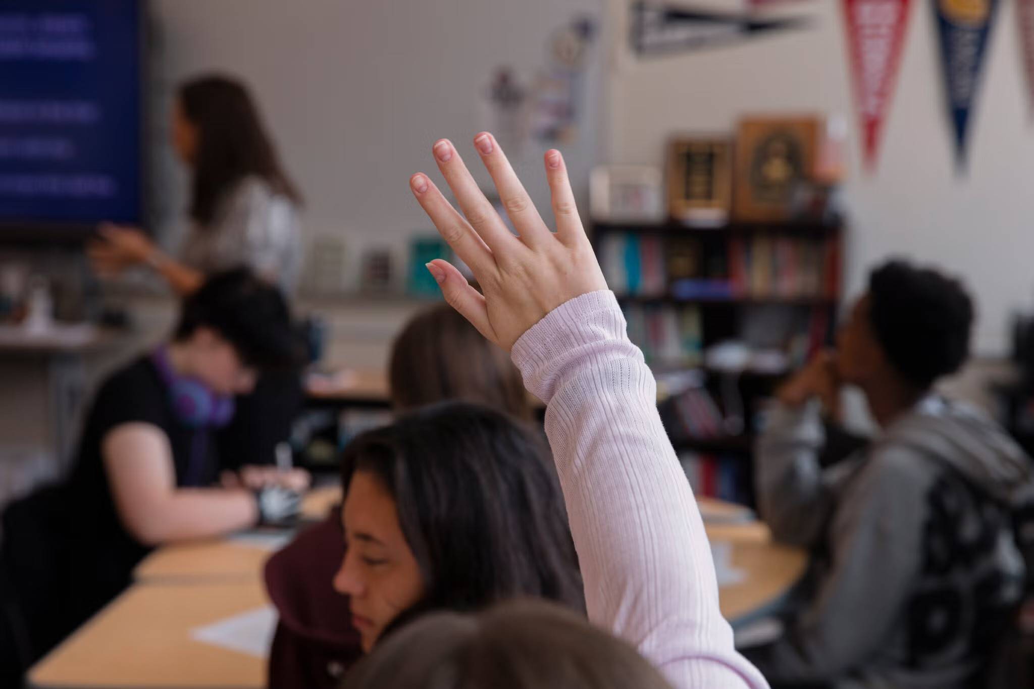 A student with arm raised in a classroom seated at a table with other students, with bookshelves and educational materials visible in the background.
