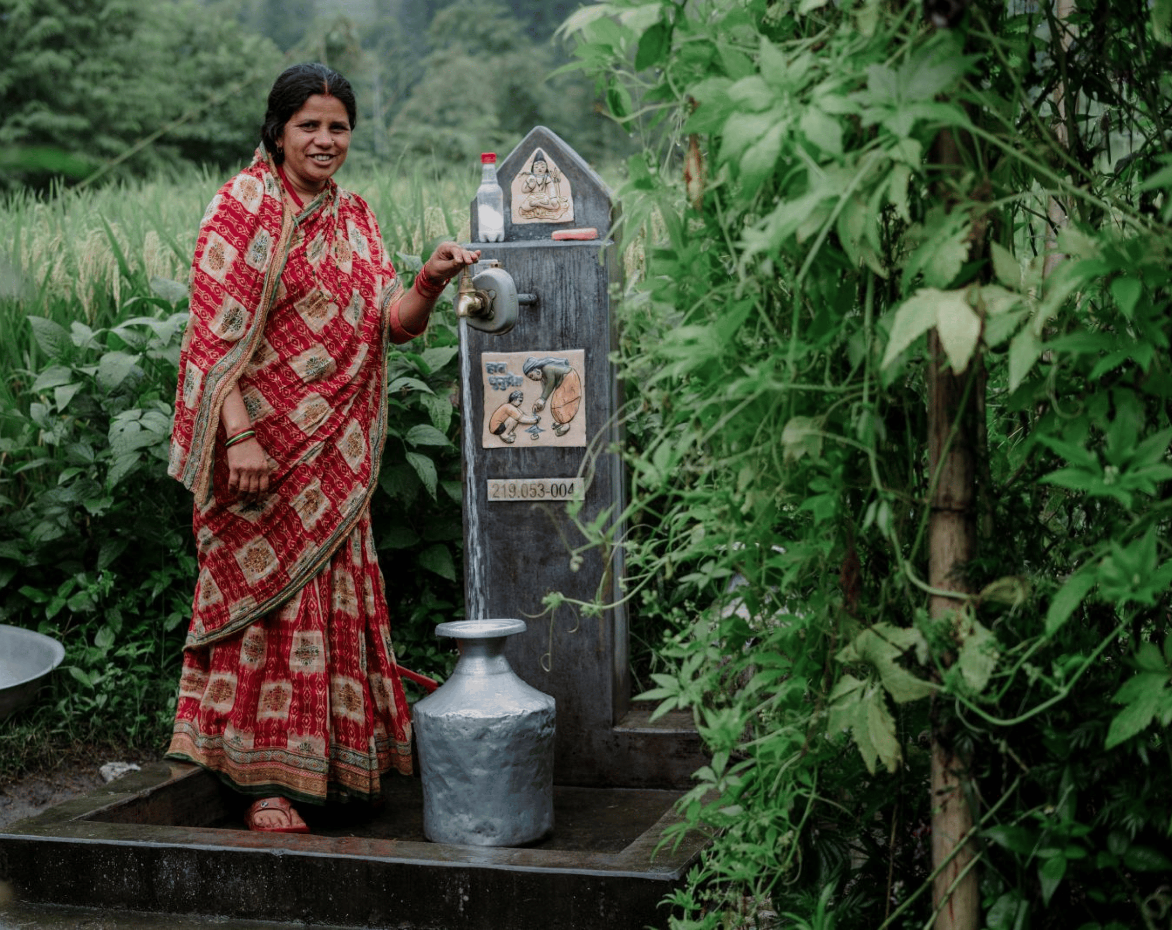 A woman in a red printed dress filling a metal water jug from a spigot surrounded by trees and foliage