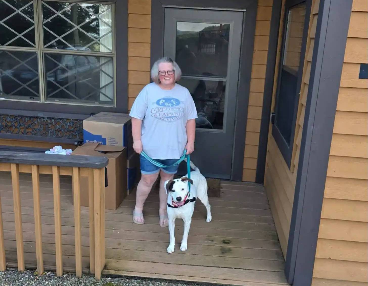 A person wearing a light blue t-shirt and glasses stands on a wooden deck with a black and white dog on a leash. Behind them is a residential structure with tan siding, a dark door, and windows.