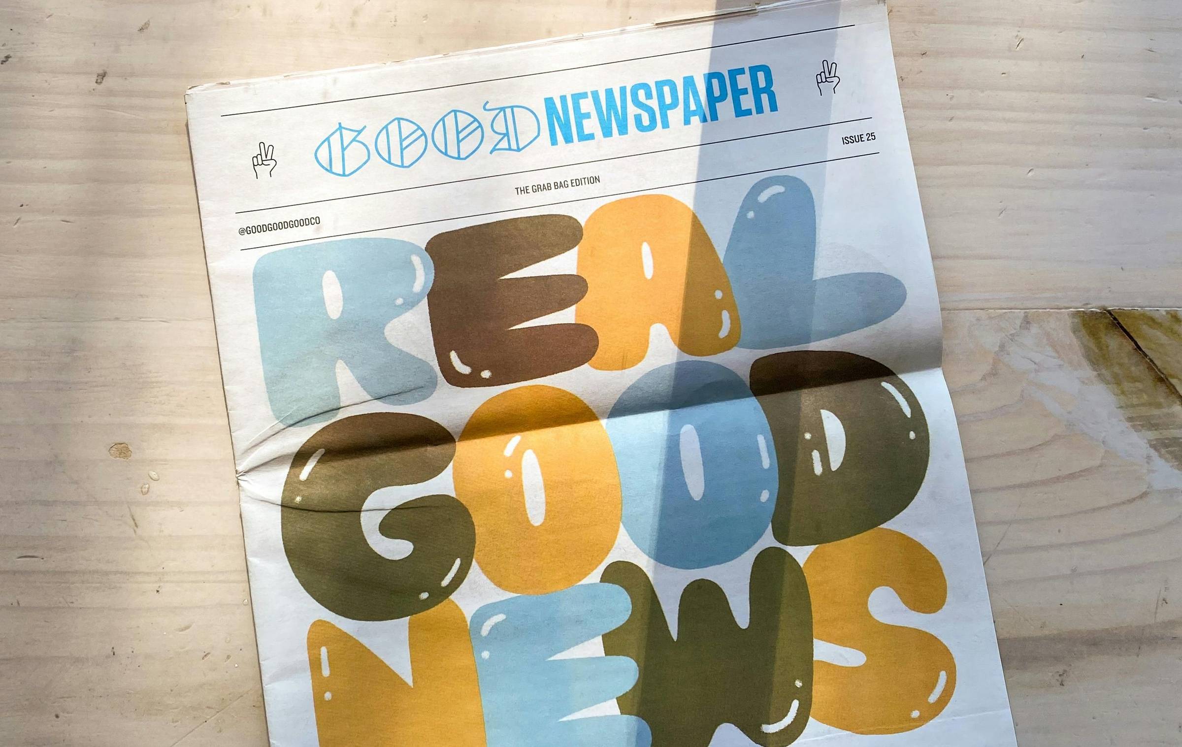 Front page of The Goodnewspaper on a wood table top, the graphic on the front says "Real Good News" in bubble letters