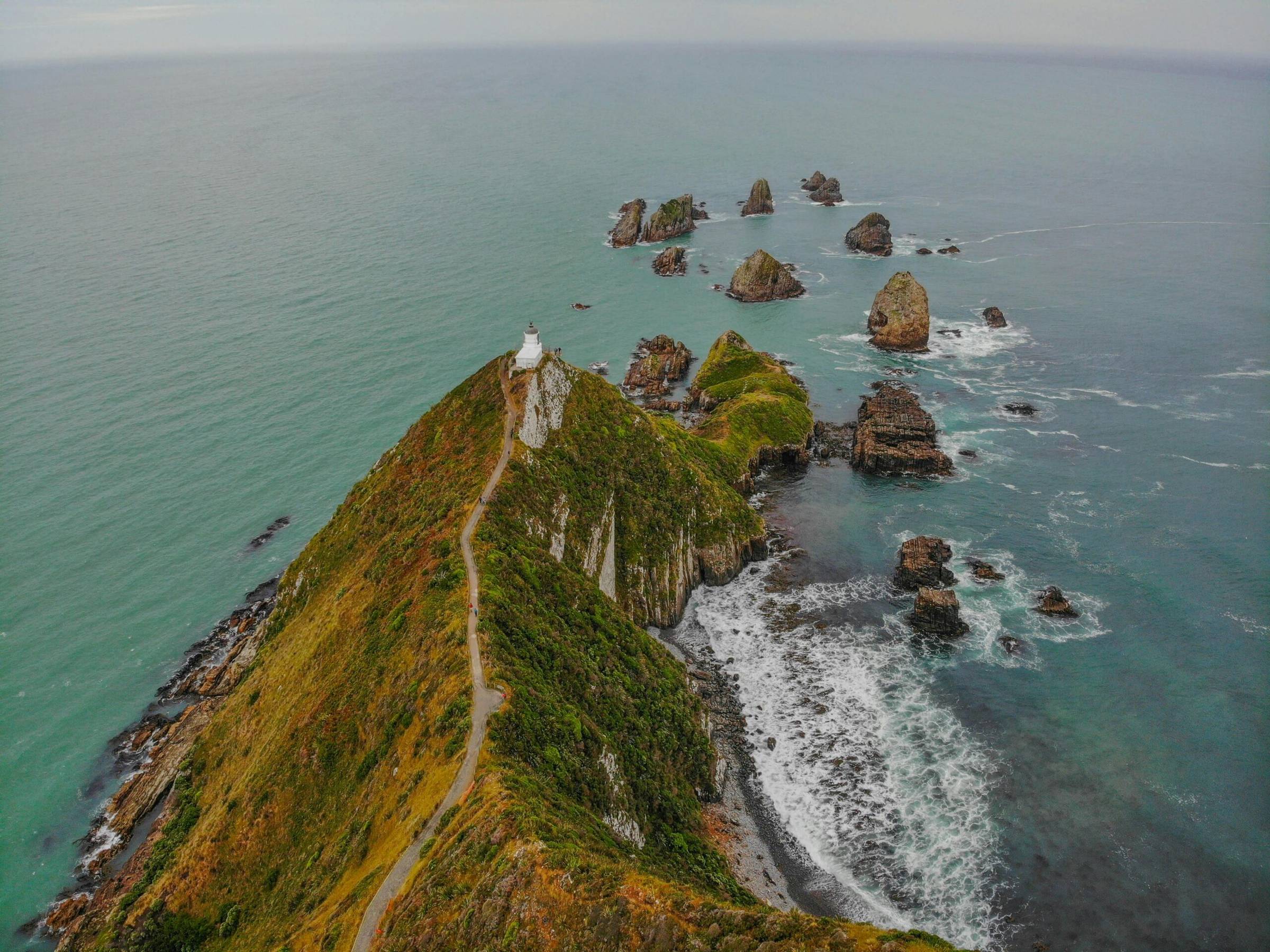 A bird's eye view of a cliffside jutting out into a waterway, there's a lighthouse at the end of it, and smaller rocks extending above the surface of the water