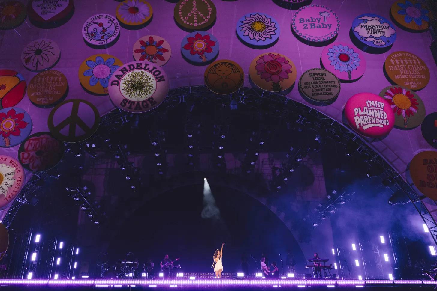 A performer stands center stage under a spotlight, surrounded by band members, with a purple-lit stage and large decorative circular buttons overhead featuring flowers, peace symbols, and text including "Daedelion Stage," "Baby 2 Baby," and "Freedom Beyon