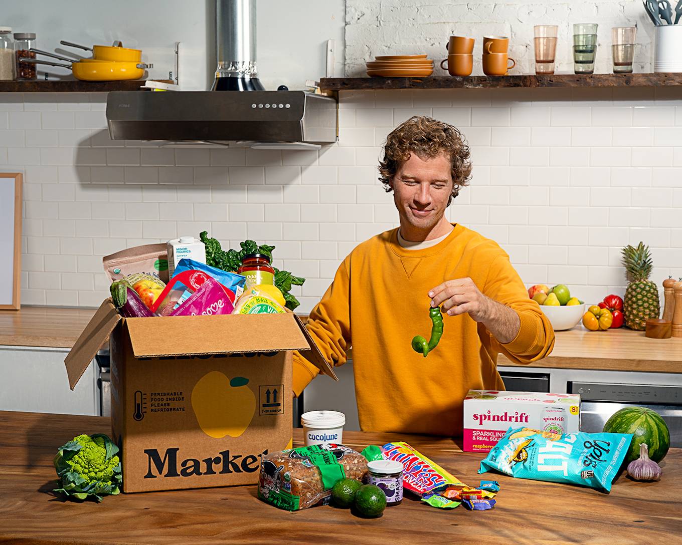 A person with light brown hair in a yellow crewneck sweatshirt standing in a kitchen next to a brown box on a counter top filled with groceries, produce and grocery items loose on the counter