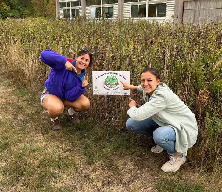 Two people pointing at a sign posted in the ground in front of a tall, grassy area