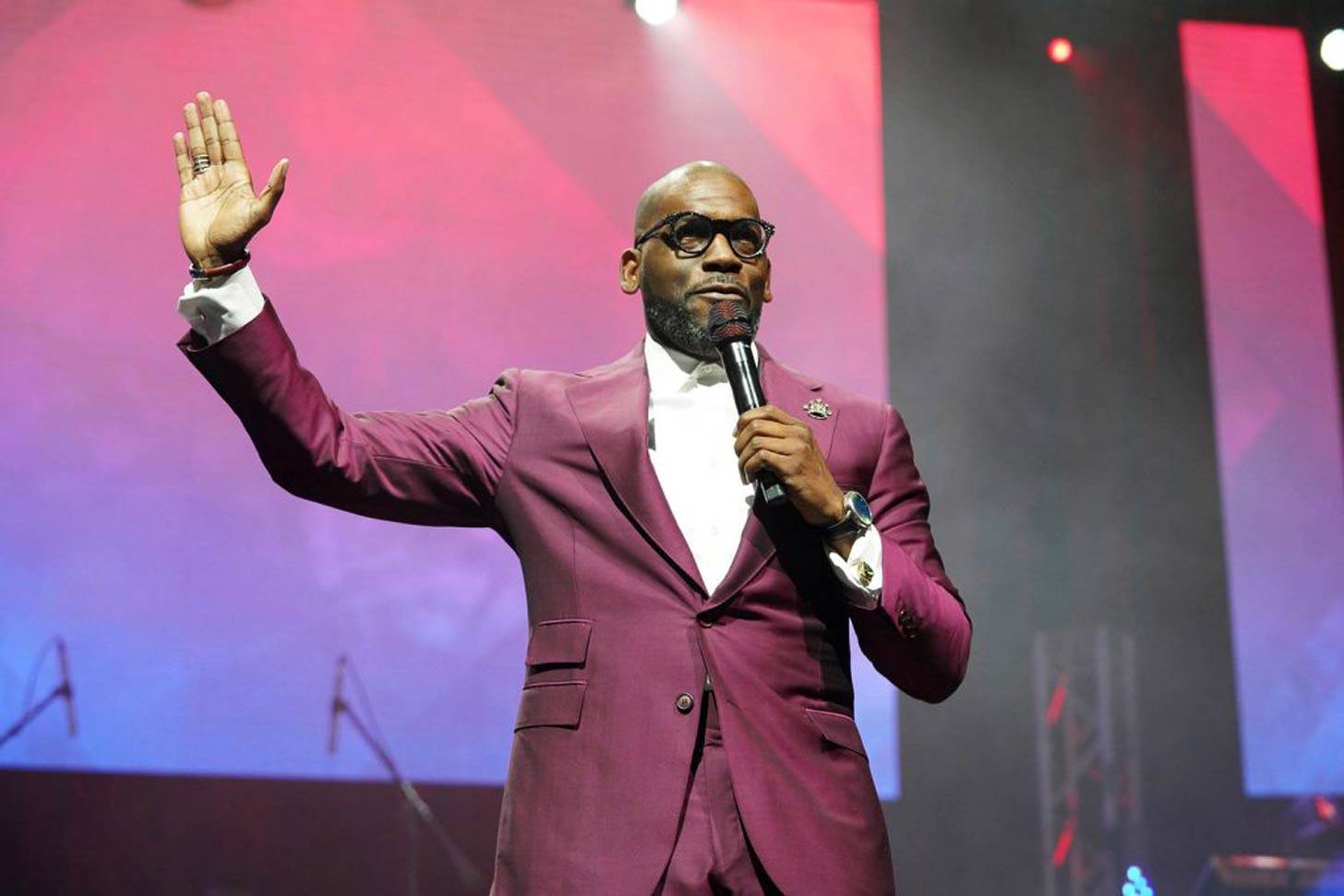 Pastor Jamal H. Bryant in a berry-colored suit holding a microphone with his right hand held up