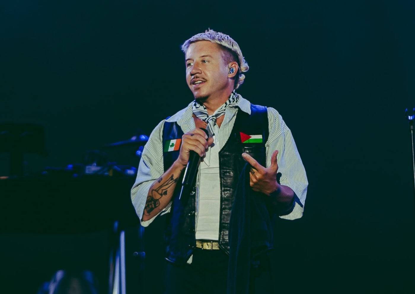 A man wearing a striped shirt and black vest holds a microphone while speaking on stage. He wears a Palestinian flag pin on his vest and has visible tattoos on his arms.