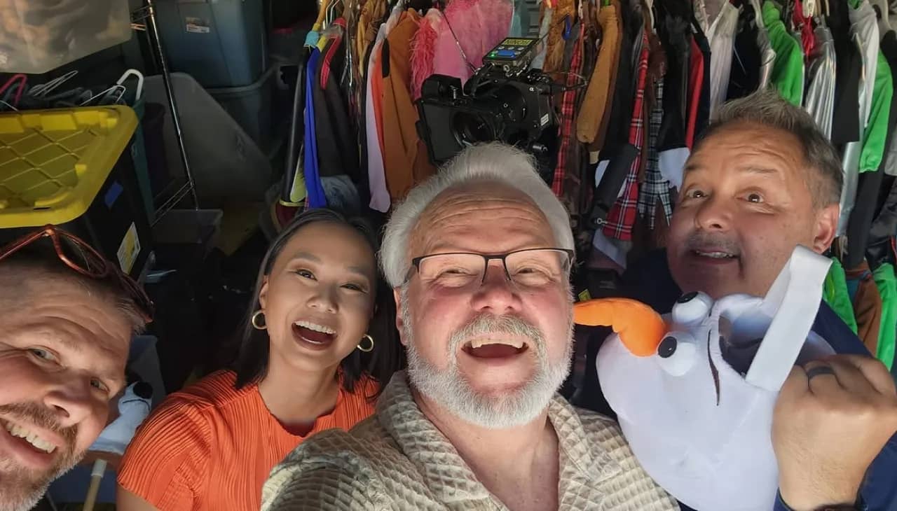 Four people smile and laugh together in a clothing storage room filled with hanging garments in various colors.