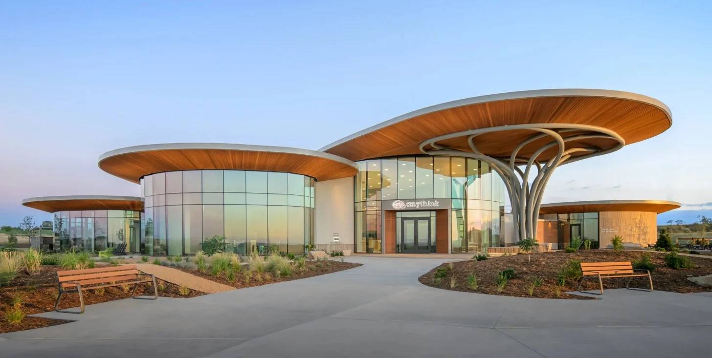 Modern building with curved wooden canopy roofs and glass walls, featuring a central entrance marked "Skylink," flanked by curved structures on either side. Concrete plaza in foreground with wooden benches and desert landscaping under clear blue sky.