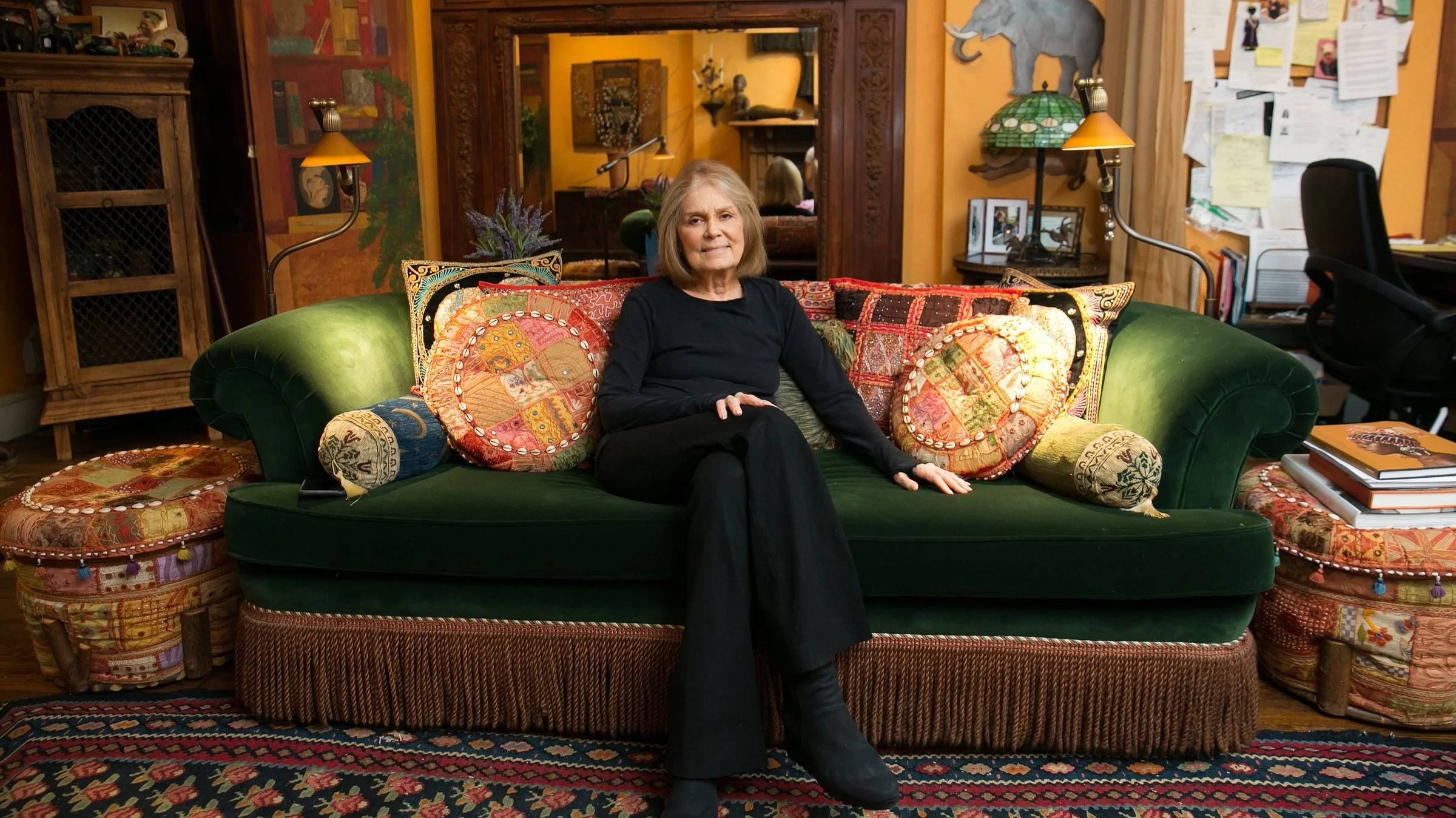 A woman in all black with short light-colored hair sitting on a green velvet sofa with colorful throw pillows