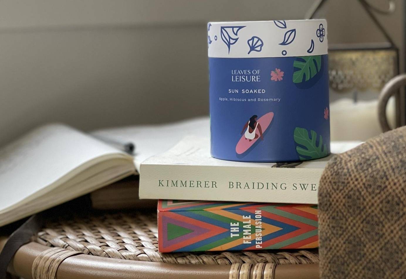 A blue, cylinder-shaped canister of "sun soaked" leaves of leisure tea sitting on top of a stack of two books