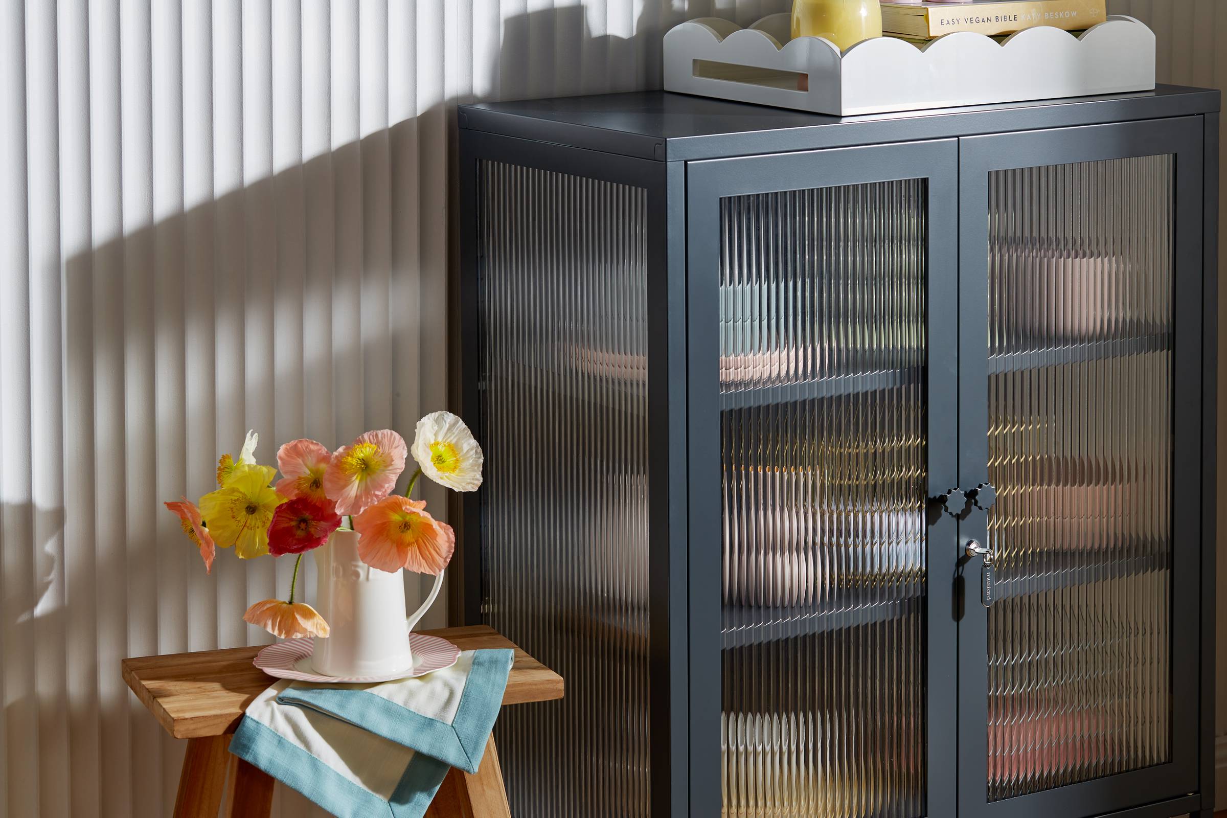 A locker-style cabinet with a black frame and fluted glass doors and sides, it's sitting in front of a scallop-textured white wall, a scalloped tray sits on top of it, and a wooden stool with a vase of flowers on top sits next to it