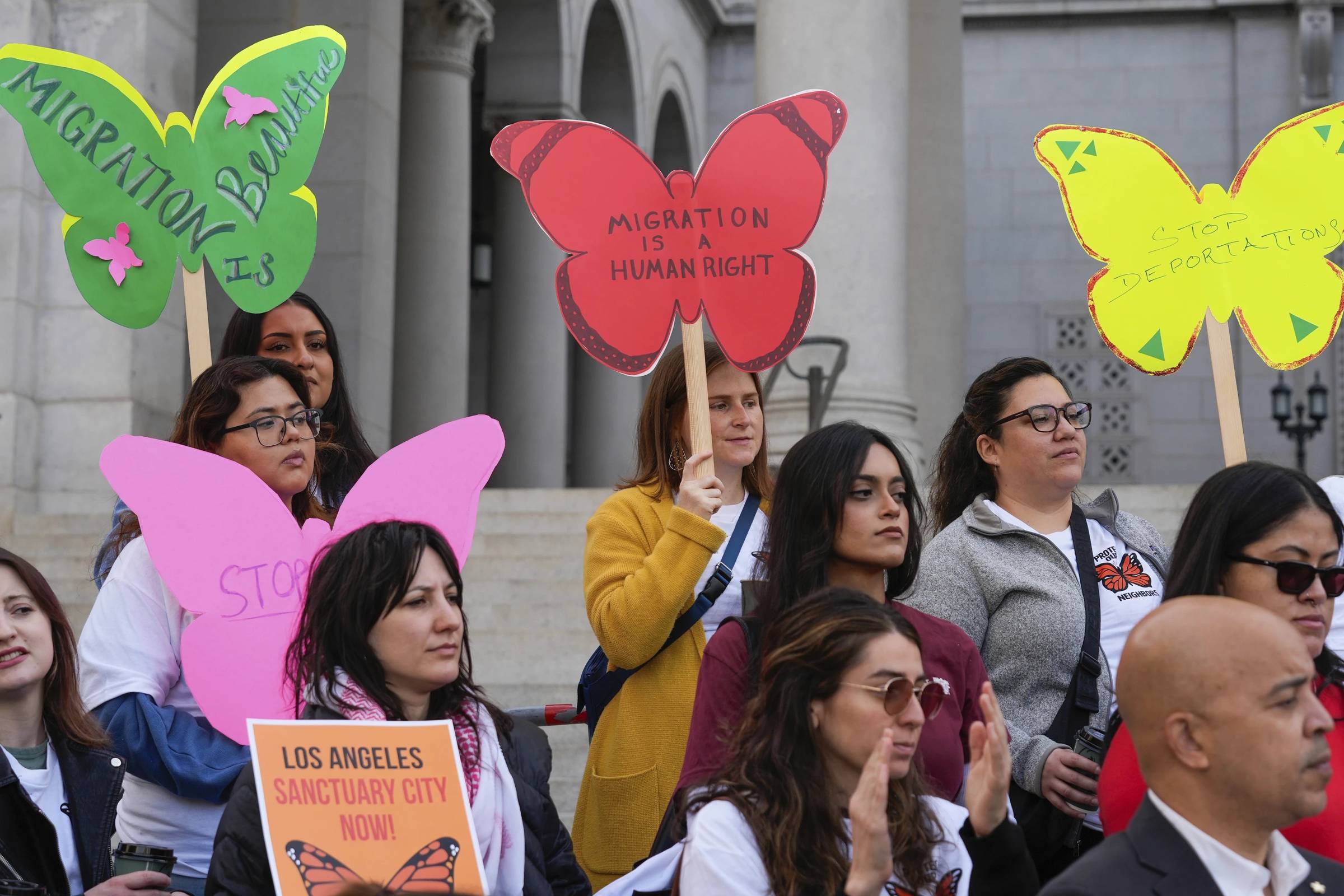 A group of people standing on steps outside a government-looking building, they're holding butterfly-shaped signs in different colors that say "Migration is beautiful" "Migration is a human right" "Stop Deportations"