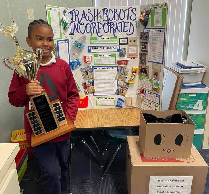 Student holding a trophy next to cardboard robot and display board