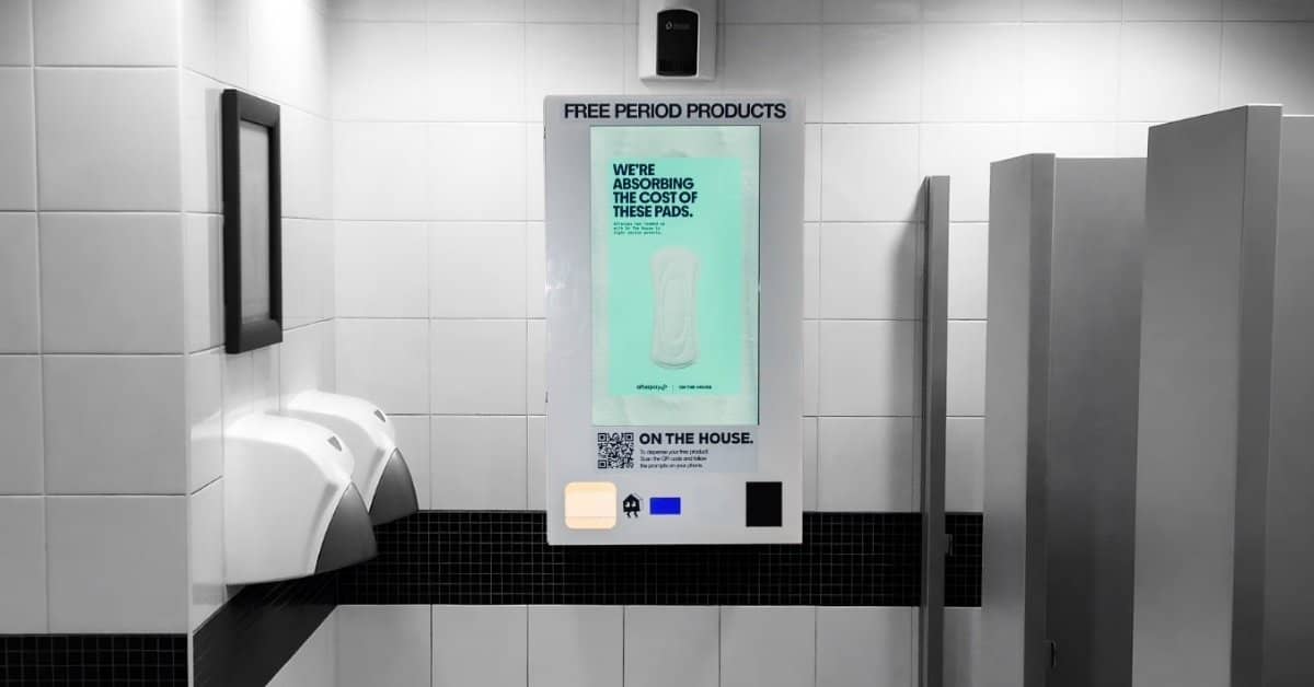 A traditional period product vending machine on a bathroom wall, this one has a screen with digital ads on it