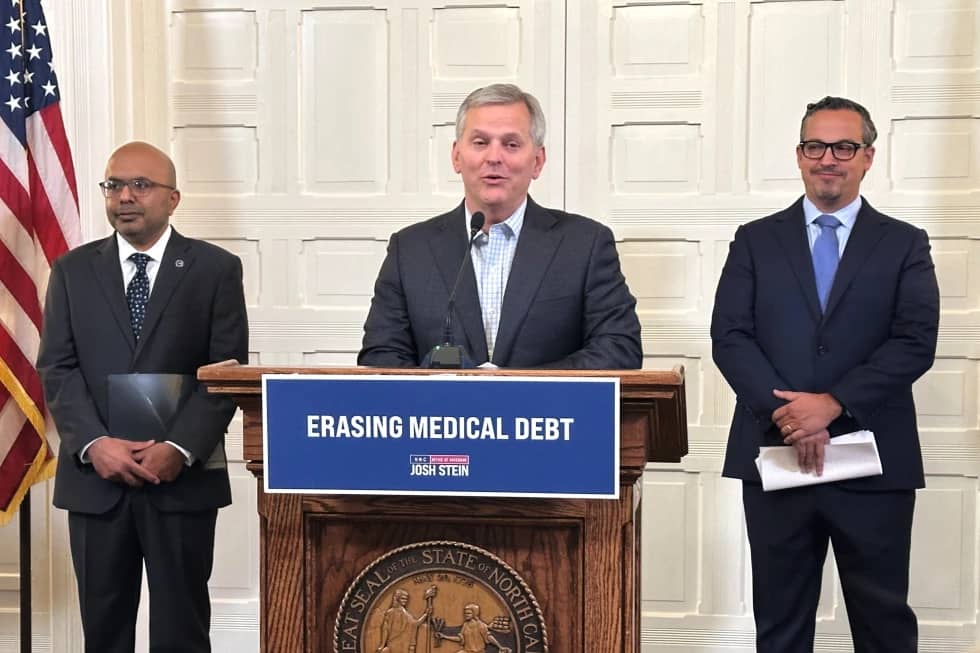 Three people standing near a podium with a sign that says "Erasing Medical Debt"