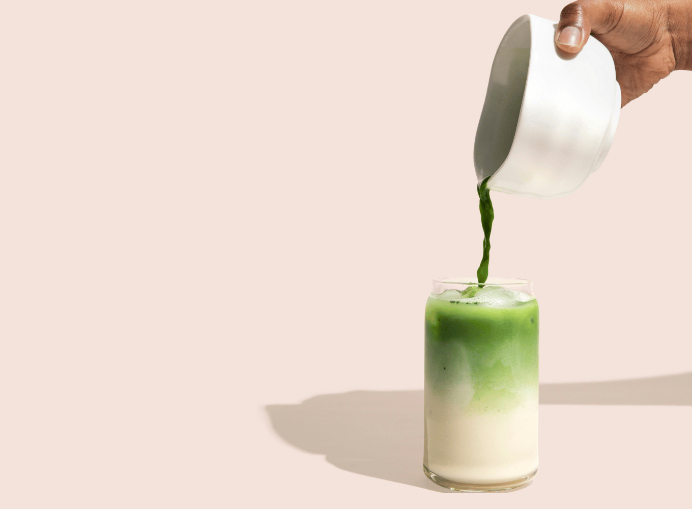 A clear glass filled with white milk with a person pouring green matcha into the top of the glass