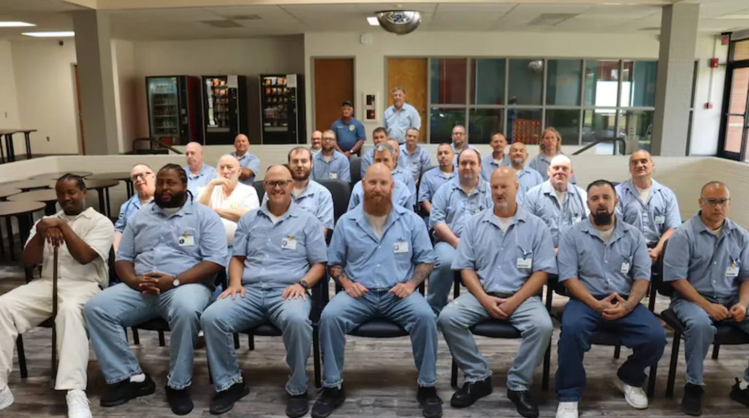 Group of men in light blue work shirts seated and standing in rows in an institutional cafeteria or break room, facing the camera.