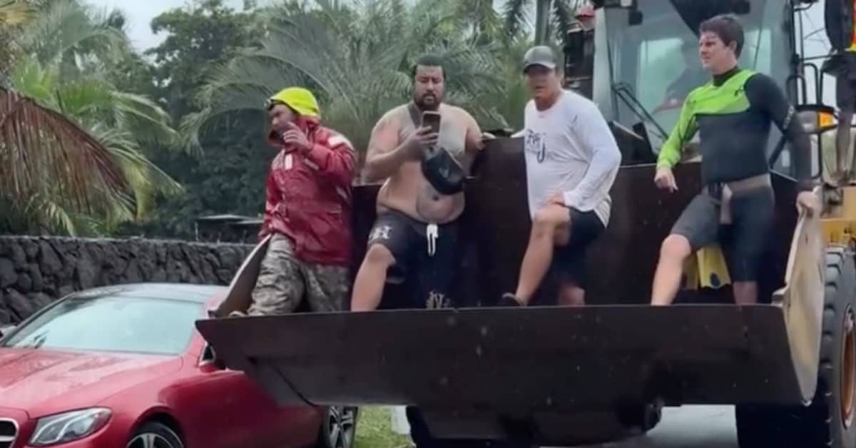 Four people standing in the bucket of a large tractor