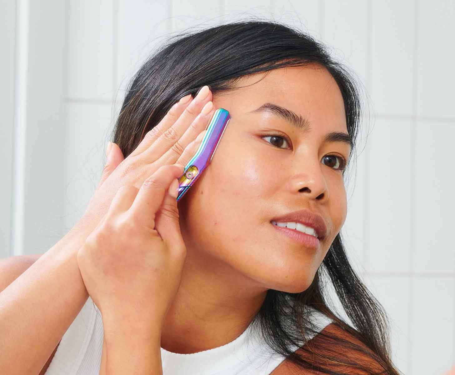 A person using a multi-colored metal Dermaplaner on their right cheek