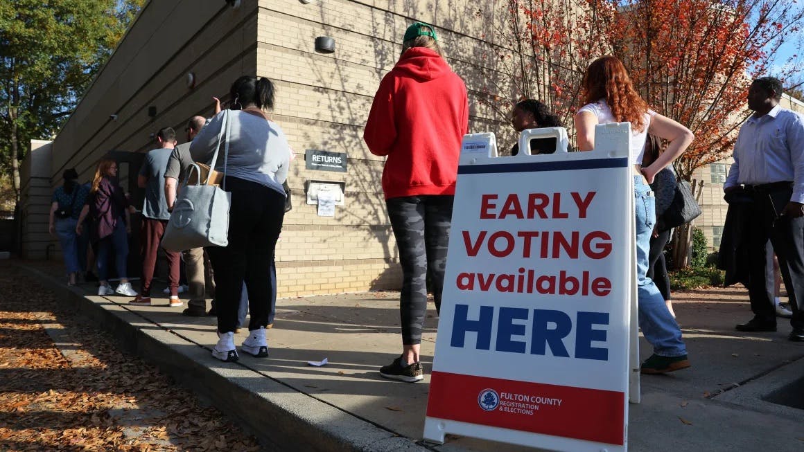 People waiting in a line outside of a building, an "Early voting available here" sandwich board sign is on the sidewalk in the foreground
